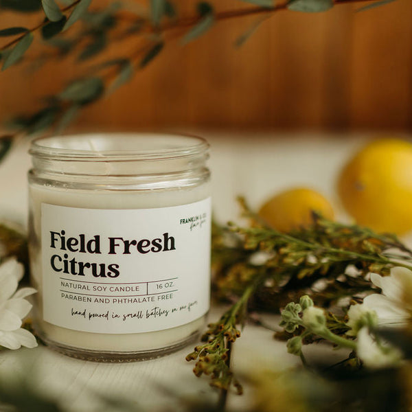 Candle labeled 'Field Fresh Citrus' surrounded by flowers and lemons on a soft background