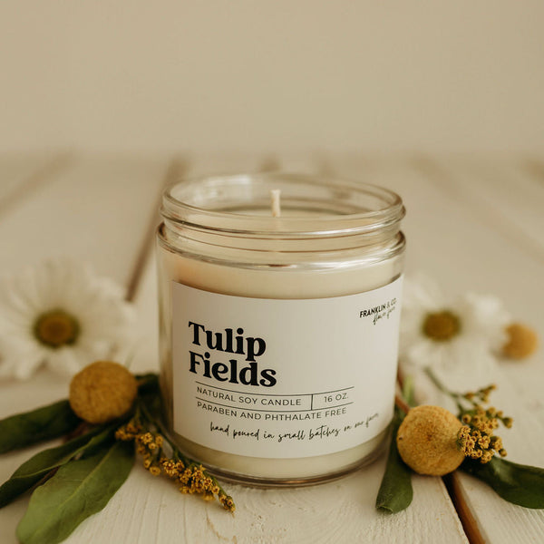 Tulip Fields candle in a glass jar with a white label on a light background with flowers.