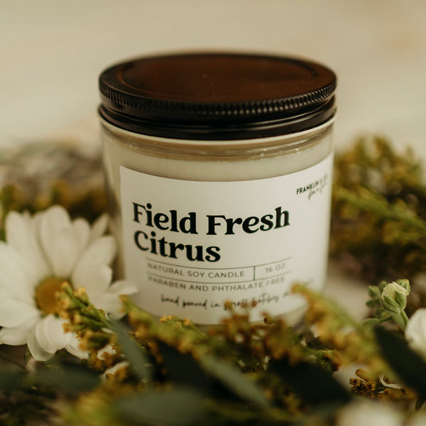 Candle labeled 'Field Fresh Citrus' surrounded by flowers on a soft background