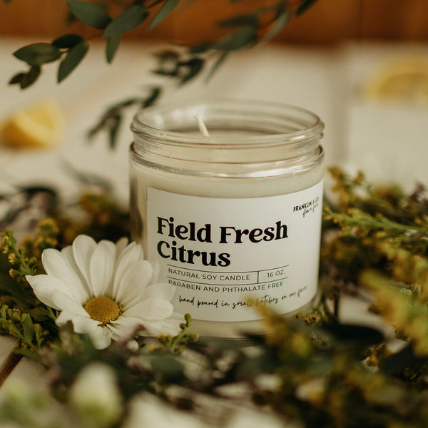 Candle in a jar labeled 'Field Fresh Citrus' surrounded by greenery and flowers.