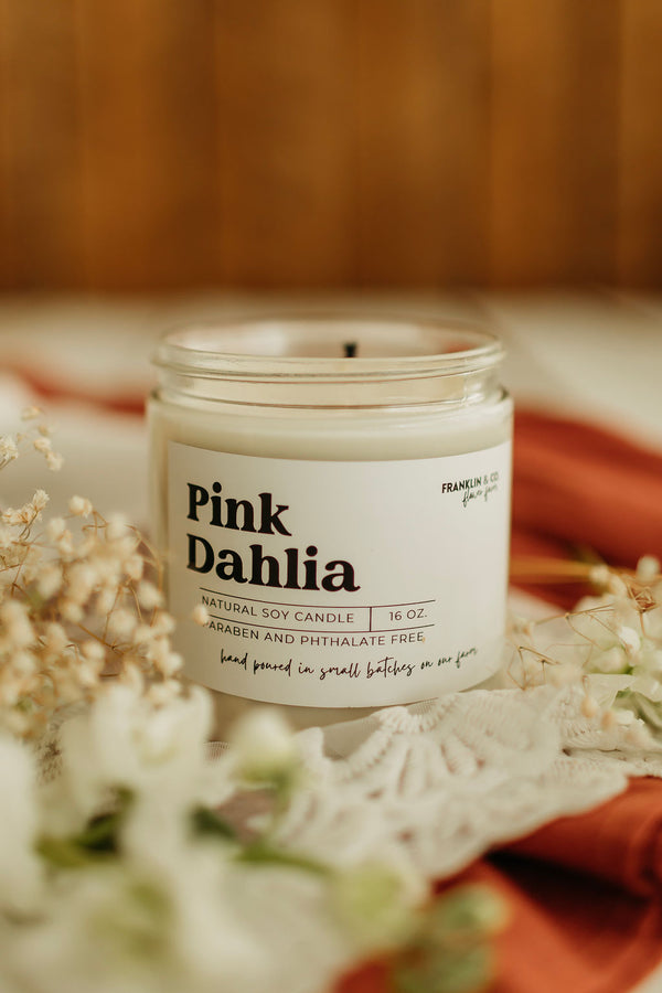Candle labeled 'Pink Dahlia' on a decorative surface with flowers and doily.