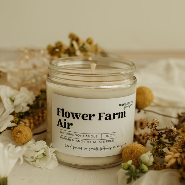 A clear glass jar soy candle with a natural cotton wick, labeled 'Flower Farm Air', surrounded by dried flowers and leaves on a white surface.