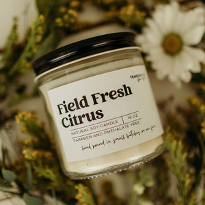 Candle labeled 'Field Fresh Citrus' surrounded by flowers on a soft background
