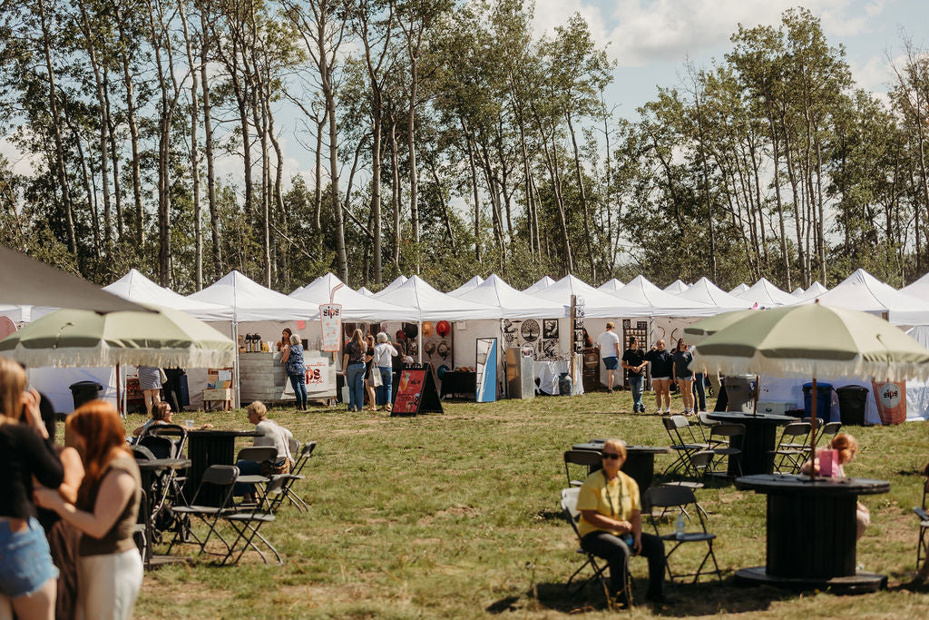 Guests relaxing at tables under umbrellas beside white vendor tents at Summer in Bloom, Charlie Lake BC