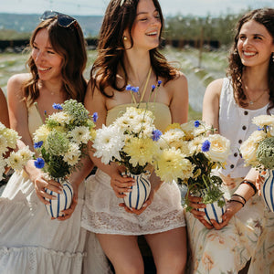 Five women sitting outdoors together, each holding a striped blue-and-white vase filled with white and yellow flowers. They are wearing light-colored dresses, and the background shows a sunny outdoor setting with greenery.