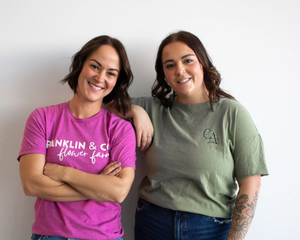 Two women standing together, one wearing a pink shirt with text and the other in a green shirt with a logo.